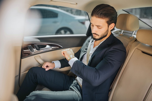 Jovem ocupado sentado no carro de luxo e olhando para os relógios. ele está atrasado. cara veste terno. ele é sério e concentrado.