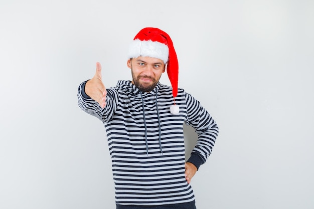 Jovem oferecendo um aperto de mão como saudação com capuz, chapéu de Papai Noel e parecendo confiante.