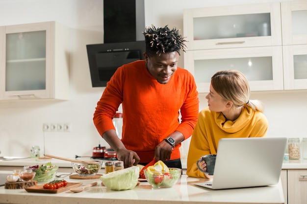 Jovem olhando atentamente para a tela de um laptop moderno enquanto corta vegetais com sua namorada curiosa em pé perto