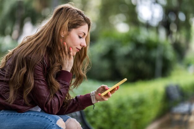 Jovem olhando para seu smartphone sentada em um banco no formato parque com espaço de cópia no perfil