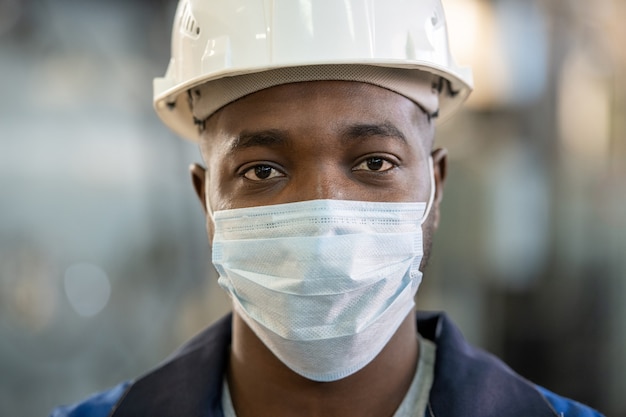 Jovem operário de etnia africana em trajes de trabalho, capacete e máscara protetora, olhando para você em pé na frente da câmera