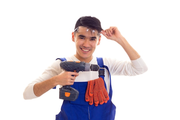 Jovem otimista com cabelo escuro na camisa branca e macacão azul com luvas laranja e óculos de proteção segurando a chave de fenda elétrica em fundo branco no estúdio.