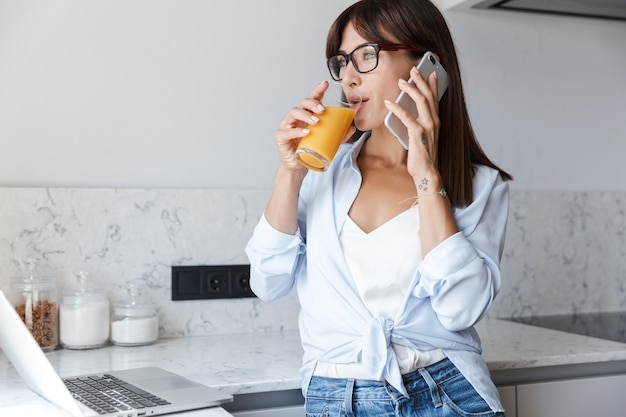jovem otimista satisfeito mulher de negócios dentro de casa em casa na cozinha usando o computador portátil, falando por telefone móvel, bebendo suco.