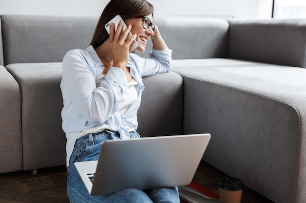 jovem otimista satisfeito mulher de negócios dentro de casa em casa, no chão, perto do sofá, usando o computador portátil, falando pelo telefone celular.