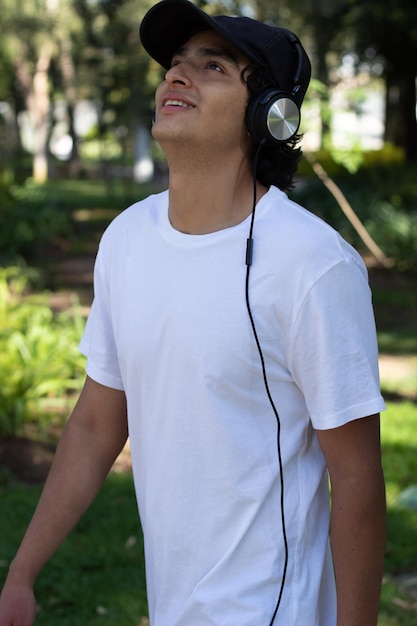 Jovem ouvindo música com fones de ouvido enquanto caminhava pelo parque sorrindo e relaxado no meio da natureza com os olhos para cima olhando para as árvores.