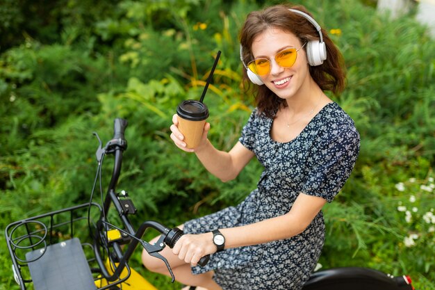 Jovem, ouvir música e andar de bicicleta no parque com uma xícara de café