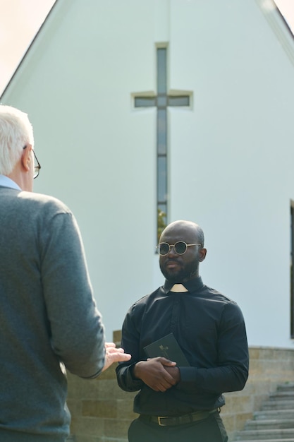 Jovem padre sério em camisa preta com colarinho clerical falando com paroquiano