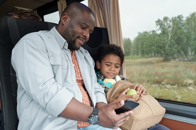 Jovem pai afro-americano sorridente usando smartphone enquanto assiste a um vídeo com o filho durante uma viagem de ônibus