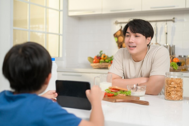 Jovem pai asiático e seu filho usando tablet digital desfrutando juntos na cozinha em casa