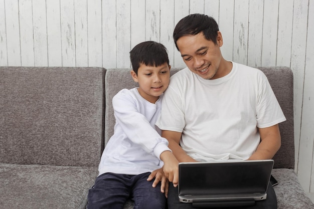 Jovem pai asiático em camiseta branca ensinando seu filho usando laptop enquanto aprende online em casa