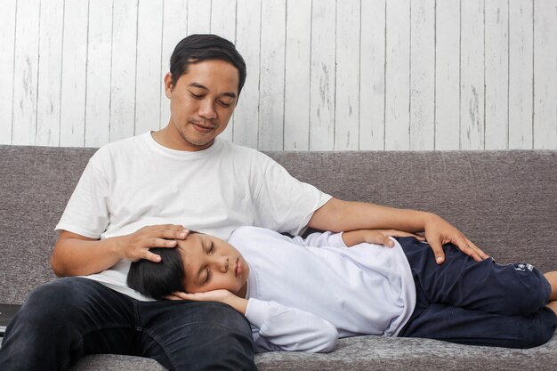 Jovem pai asiático em camiseta branca está acariciando a cabeça de seu filho enquanto dorme no sofá