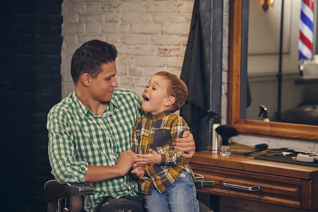 Foto jovem pai bonito e seu filho pequeno e elegante na barbearia esperando o barbeiro. o pai está sentado na cadeira e o filho está perto