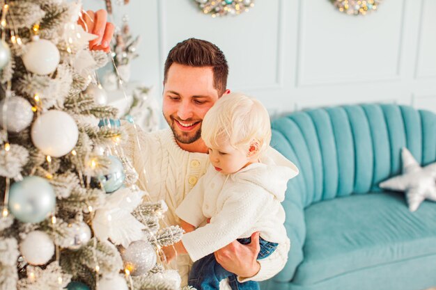 Jovem pai bonito segurando seu filho adorável com cabelo loiro por um lado e pendurando brinquedos de natal na árvore com outro menino bonitinho olhando para a árvore
