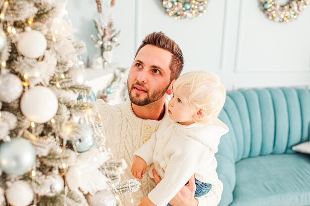 Jovem pai bonito segurando seu filho adorável com cabelo loiro por um lado e pendurando brinquedos de natal na árvore com outro menino bonitinho olhando para a árvore