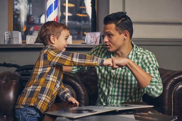 Foto jovem pai e seu filho elegante na barbearia na sala de espera. eles estão esperando o mestre, lendo a revista de moda e se divertindo