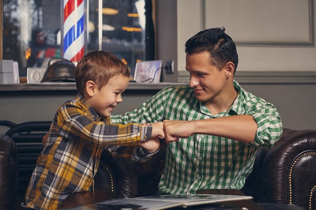 Foto jovem pai e seu filho elegante na barbearia na sala de espera. eles estão esperando o mestre, lendo a revista de moda e se divertindo