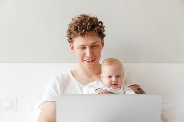 Jovem pai feliz brincando com seu filho bebê enquanto trabalhava em um laptop, deitado na cama