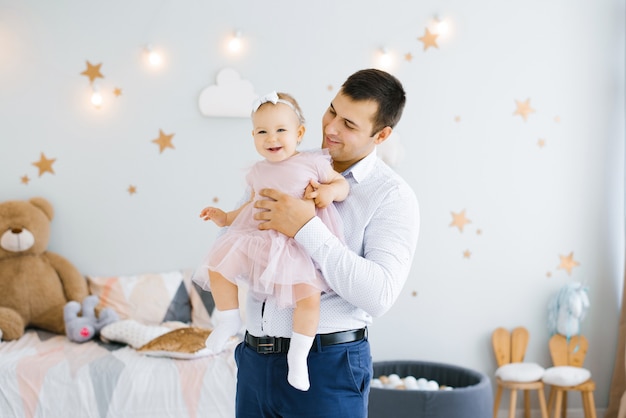 Jovem pai feliz com uma filha sorridente de um ano nos braços no quarto das crianças