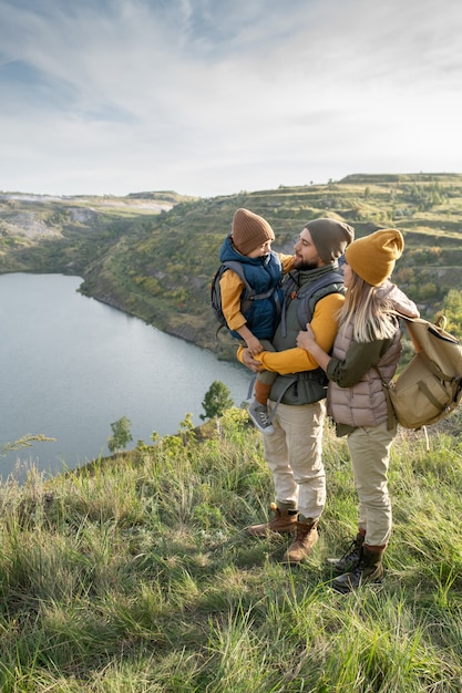 Jovem pai segurando o filho nas mãos com uma mulher afetuosa em pé ao lado deles contra um lago ou rio cercado por montanhas durante a viagem