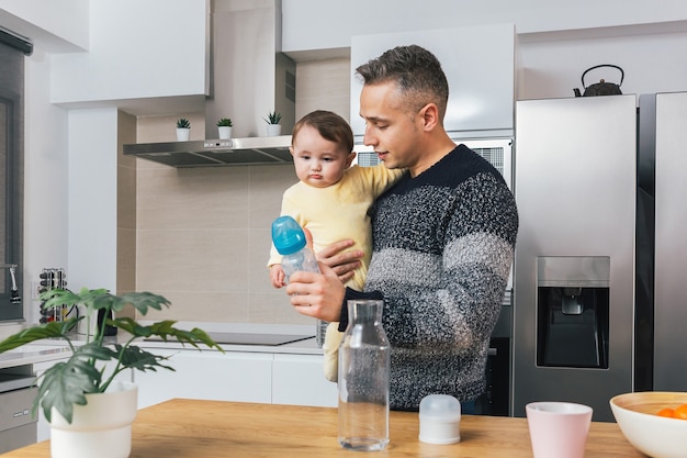 Foto jovem pai solteiro preparando uma mamadeira enquanto segura seu bebê nos braços em casa