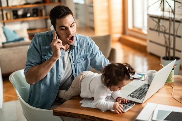 Jovem pai trabalhador falando ao telefone enquanto cuidava de sua filha pequena em casa