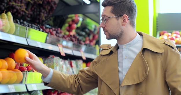 Jovem parado no supermercado e escolhendo laranjas Homem estiloso olhando frutas enquanto faz compras