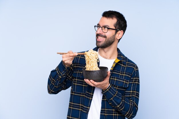 Jovem, parede azul, segurando uma tigela de macarrão com pauzinhos e olhando para cima