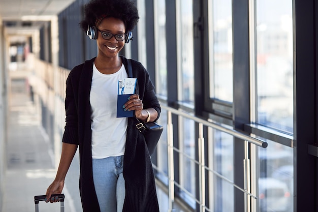 Jovem passageira afro-americana em roupas casuais e fones de ouvido está no aeroporto com bagagem.