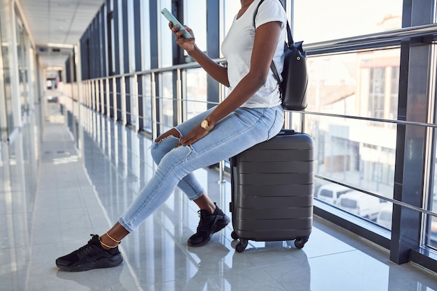 Jovem passageira afro-americana em roupas casuais está no aeroporto com bagagem. Usando telefone.