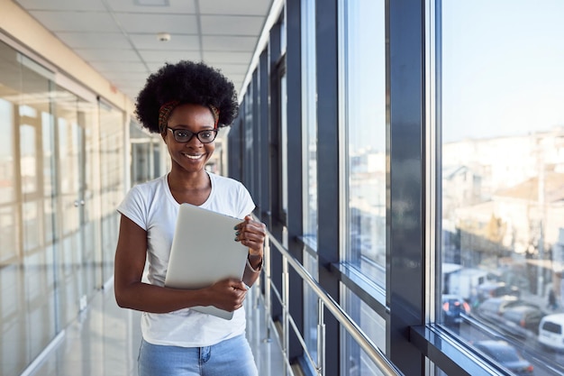 Jovem passageira afro-americana em roupas casuais está no aeroporto segurando laptop.