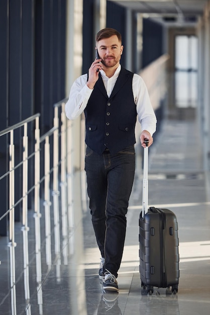 Jovem passageiro masculino em roupas formais elegantes está no saguão do aeroporto andando com bagagem e falando por telefone.
