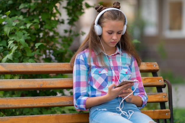 Jovem pensativa selecionando uma nova trilha sonora em seu celular enquanto se senta em um banco ao ar livre ouvindo música
