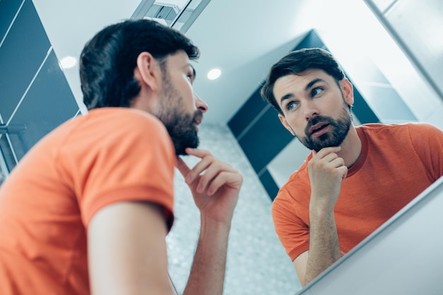 Foto jovem pensativo e curioso em pé na frente de um espelho e tocando seu queixo enquanto olha para o reflexo