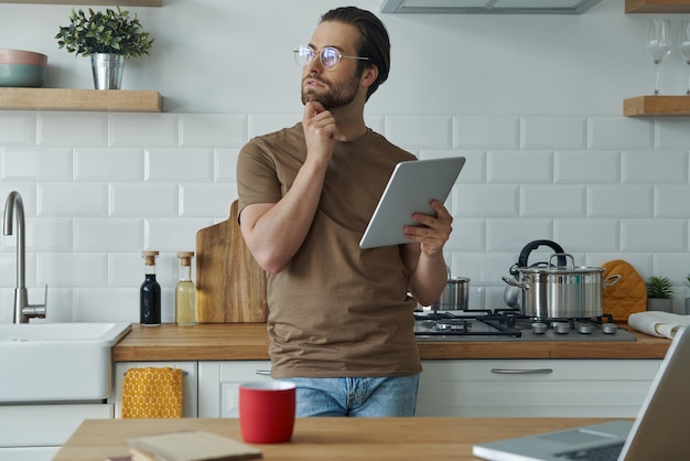 Jovem pensativo usando tecnologias enquanto trabalhava na cozinha doméstica