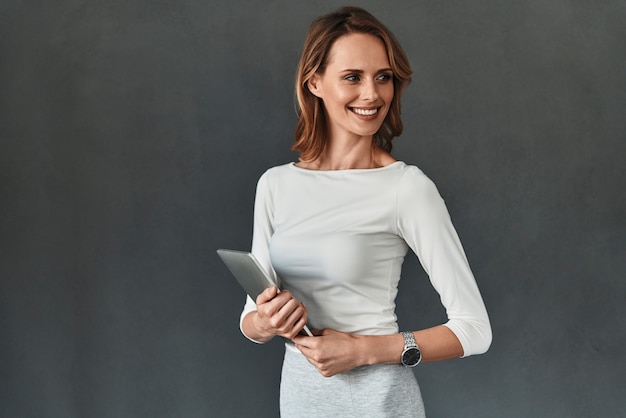 Jovem perfeccionista. Mulher jovem e bonita em smart casual wear segurando um tablet digital e sorrindo em pé contra um fundo cinza