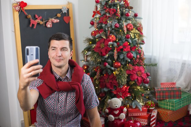 Jovem perto da árvore de natal em casa com roupas confortáveis acena com a mão em sinal de olá / adeus na frente da tela do smartphone. parabéns pela conexão de vídeo com o natal e ano novo.