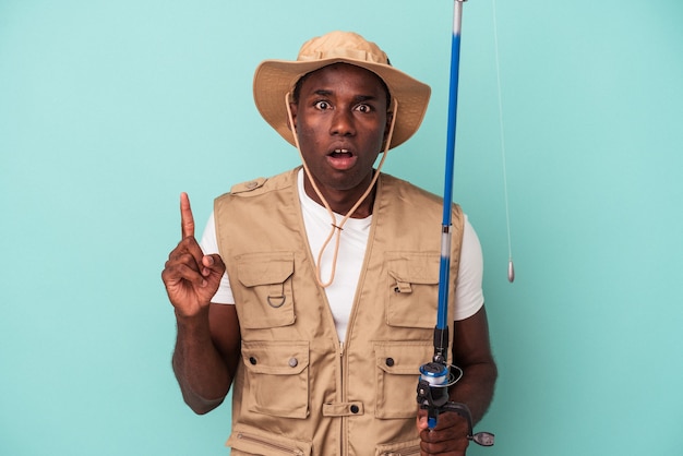 Jovem pescador afro-americano segurando a haste isolada no fundo azul, tendo uma ideia, o conceito de inspiração.