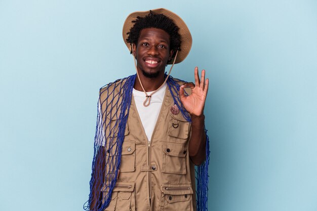 Jovem pescador afro-americano segurando uma rede isolada em um fundo azul alegre e confiante, mostrando um gesto ok.