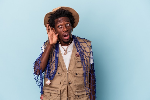 Jovem pescador afro-americano segurando uma rede isolada em um fundo azul, tentando ouvir uma fofoca.