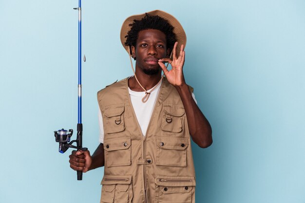 Jovem pescador afro-americano segurando uma vara isolada em um fundo azul com os dedos nos lábios, mantendo um segredo.