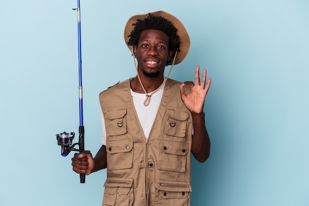 Jovem pescador afro-americano segurando uma vara isolada no fundo azul, alegre e confiante, mostrando o gesto de ok.