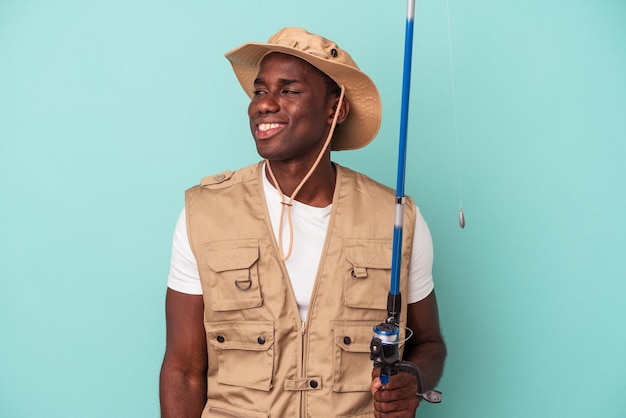 Jovem pescador afro-americano segurando vara isolada em fundo azul olha de lado sorrindo alegre e agradável