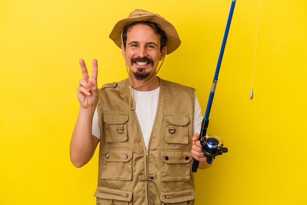 Jovem pescador caucasiano segurando a haste isolada em um fundo amarelo, mostrando o número dois com os dedos.