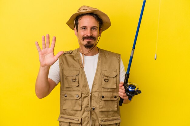 Jovem pescador caucasiano segurando a haste isolada no fundo amarelo, sorrindo alegre mostrando o número cinco com os dedos.