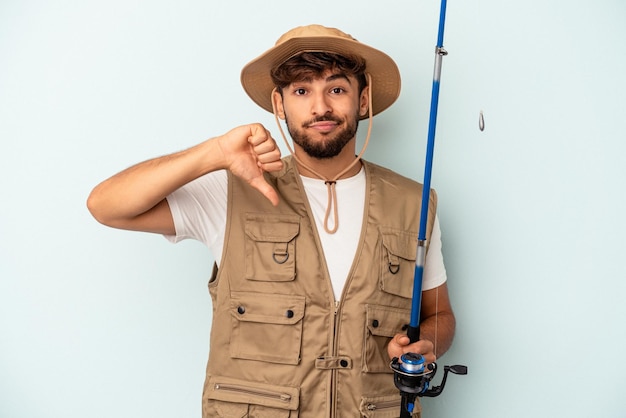 Jovem pescador de raça mista segurando uma vara isolada em um fundo azul, mostrando um gesto de antipatia, polegares para baixo. Conceito de desacordo.