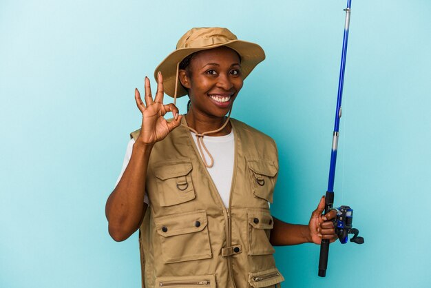 Jovem pescadora afro-americana segurando a haste isolada no fundo azul, alegre e confiante, mostrando o gesto de ok.