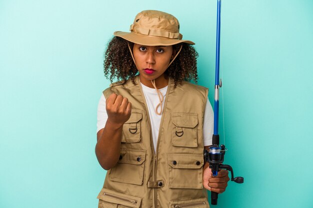 Jovem pescadora afro-americana segurando uma vara isolada em um fundo azul, mostrando o punho para a câmera, expressão facial agressiva.