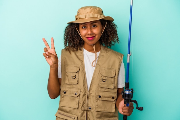 Jovem pescadora afro-americana segurando uma vara isolada sobre fundo azul, mostrando o número dois com os dedos.