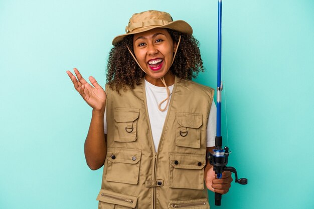 Jovem pescadora afro-americana segurando uma vara isolada sobre fundo azul, recebendo uma agradável surpresa, animada e levantando as mãos.