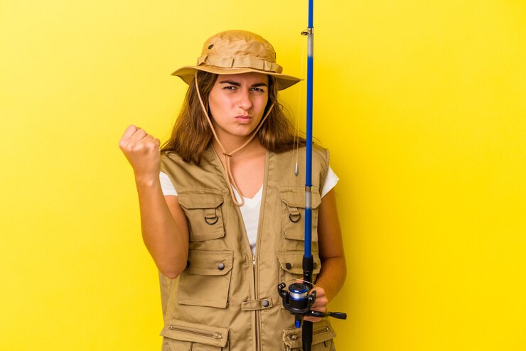 Foto jovem pescadora caucasiana segurando uma haste isolada em fundo amarelo, mostrando o punho para a câmera, expressão facial agressiva.